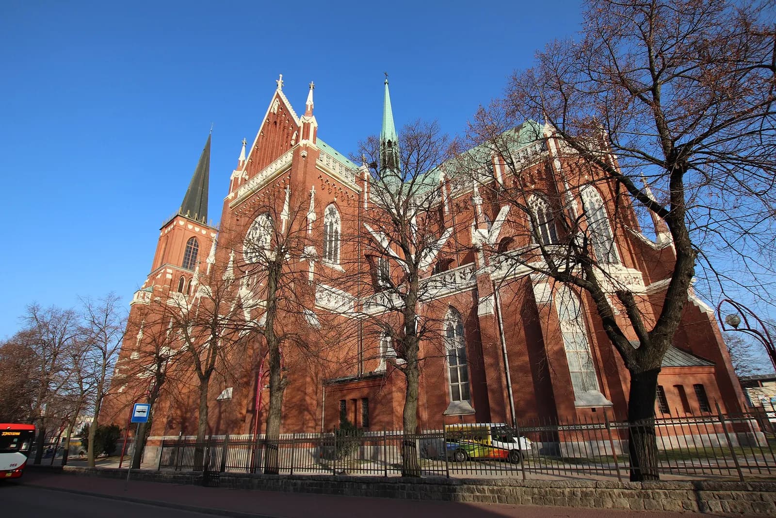 A large red brick church with a green roof and a prominent spire stands under a clear blue sky, partially obscured by bare winter trees.