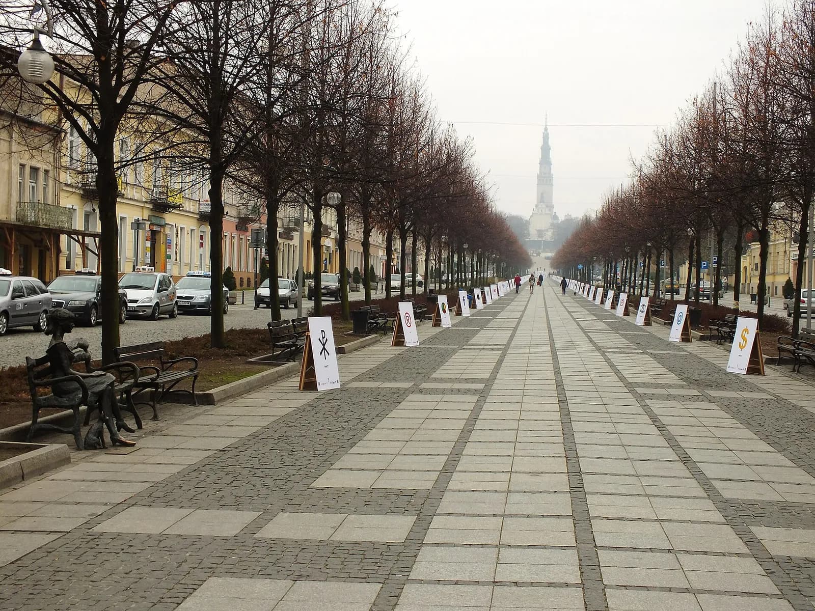 A long, tree-lined pedestrian street stretches towards a distant church spire on an overcast day, with buildings and parked cars on the left.